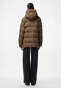 Chaqueta acolchada marrón con capucha, con un diseño acolchado y un ajuste relajado. Combinada con pantalones anchos oscuros. Sin herrajes visibles.