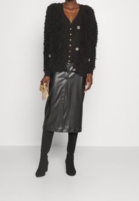 Schwarze, flauschige Strickjacke mit goldenen Knöpfen, kombiniert mit einem schwarzen Leder-Bleistiftrock und kniehohen, schwarzen Stiefeln mit Absatz für einen eleganten Look.