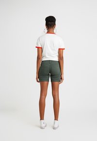 Camiseta blanca con ribetes rojos, pantalones cortos verdes y zapatillas blancas. Bolsillos traseros visibles en los pantalones cortos, textura lisa y diseño casual.
