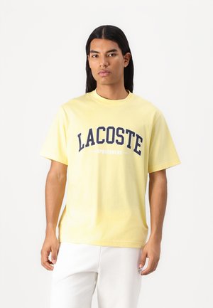 Geltona medvilninė marškinėliai su trumpomis rankovėmis, ant priekio turinti tamsiai mėlyną "LACOSTE SPORTSWEAR" logotipą.
