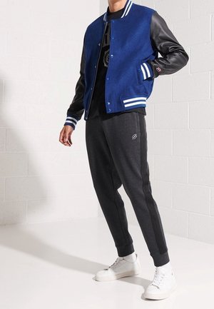 Veste varsity bleue avec manches en cuir noir, rayures blanches, et un chemisier noir en dessous. Pantalon de survêtement gris avec rayure latérale et baskets blanches.