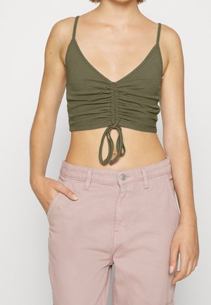 Femme portant un crop top froncé vert olive à fines bretelles et un pantalon taille haute rose pâle, une main dans la poche.