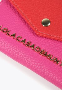 Cartera de cuero texturizado rosa con solapa roja, herrajes dorados y logo en relieve "LOLA CASADEMUNT". Presenta un acabado suave y un diseño estructurado.