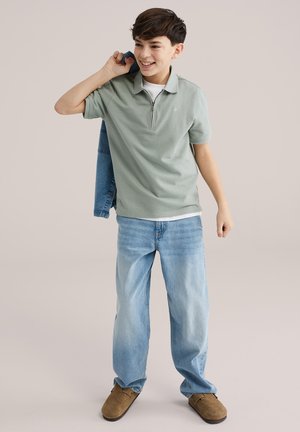 Fant v zeleni polo majici, svetlo modrih kavbojkah in rjavih čevljih, ki drži jeans jakno preko ramena in se smehlja.