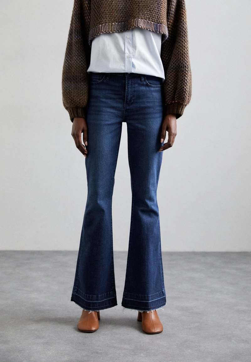 Jeans in denim scuro a zampa d'elefante con orlo grezzo, abbinati a un maglione corto marrone a trama e a una camicia azzurra con colletto. Mules con tacco marroni.
