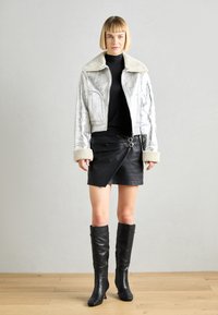 Chaqueta de cuero sintético plateada con cuello de piel, suéter negro de cuello alto, falda de cuero con detalles de hebilla y botas negras de tacón alto hasta la rodilla.