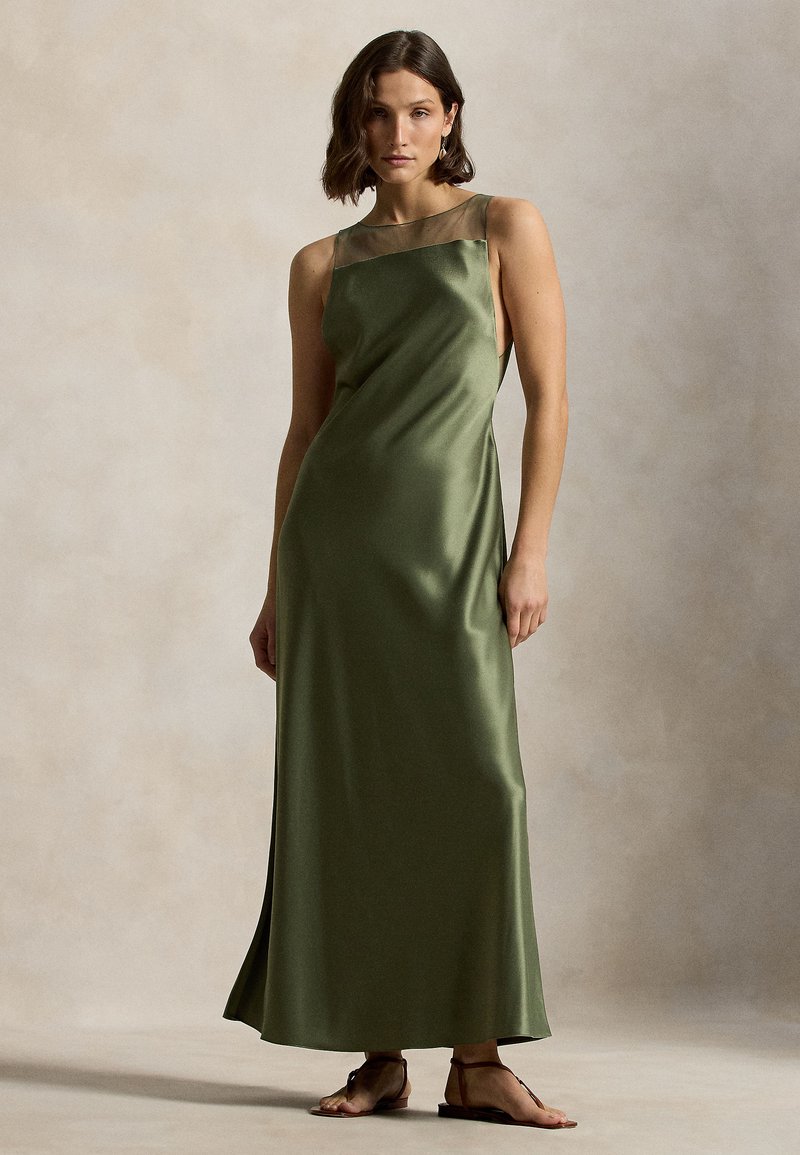 Oliivinvihreä satiininen maksimekko, jossa on neliskanttinen pääntie ja ohuet hihnat, sileä pinta ja sivuhalkiot. Päällä yksinkertaiset ruskeat tasapohjaiset sandaalit.