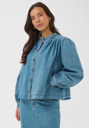 Femme aux longs cheveux foncés portant une veste en jean bleue boutonnée avec des manches bouffantes et une jupe en jean assortie sur un fond uni.