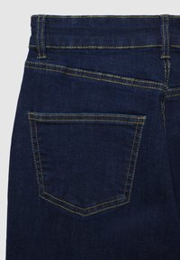 Jeans in denim blu scuro con una vita a metà altezza, caratterizzati da una tasca posteriore bordata da cuciture gialle e una texture liscia.