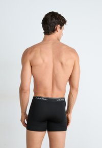 Boxeri negri dintr-un material neted, cu o croială strânsă. Banda elastică are un logo alb Calvin Klein pe un fundal negru uniform.