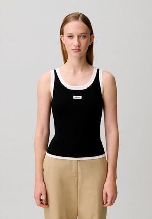 Joven mujer con el pelo liso que lleva una camiseta negra sin mangas con ribete blanco y pantalones beige sobre un fondo claro y liso.