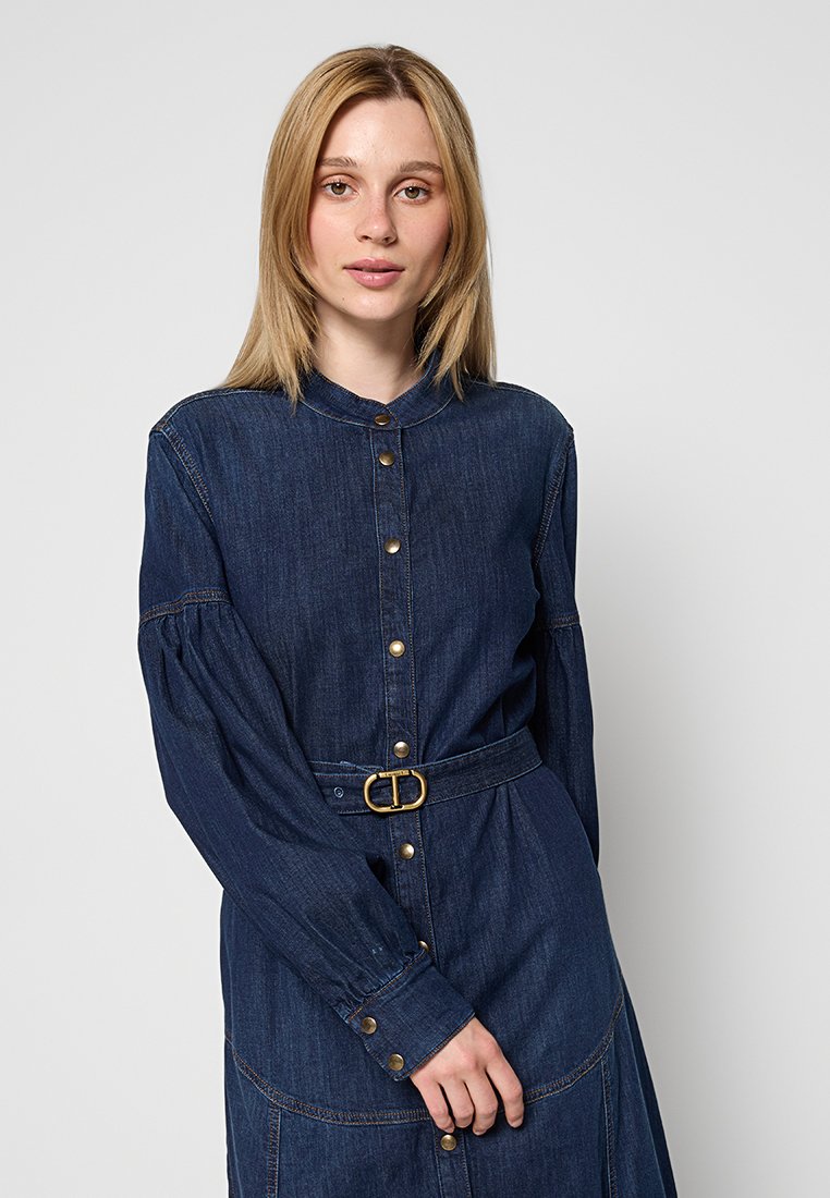 Dżinsowa sukienka z kołnierzykiem, długimi bufkami, z przodu na guziki oraz z paskiem w talii. Ciemnoniebieski kolor, gładka faktura i złote guziki.