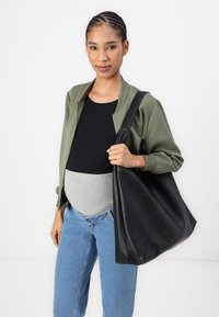 Borsa tote in pelle nera con una superficie liscia, dotata di una sola tracolla, abbinata a una giacca bomber verde oliva, top nero e jeans blu.