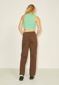 Calças castanhas de perna reta com dois bolsos traseiros, usadas com um top cropped verde. Textura de algodão suave e design simples. Sapatos pretos completam o visual.