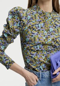 Top floreale con maniche a sbuffo in tonalità di verde, viola e blu. Collo rotondo, abbinato a una borsa a tracolla viola e jeans.
