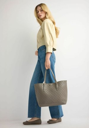 Kobieta o długich blond włosach, ubrana w beżową bluzkę, niebieskie dżinsy i brązowe baleriny, trzymająca obok siebie dużą, pikowaną torbę w kolorze taupe.