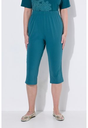 Mujer con pantalones recortados color azul verdoso y top a juego con diseño de flor de crochet, combinados con zapatos blancos sin cordones con estampado floral, de pie contra una pared blanca.