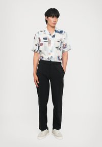 Camisa de botón de manga corta en blanco con patrones abstractos coloridos; combinada con pantalones negros y zapatillas blancas, de pie con las manos en los bolsillos.