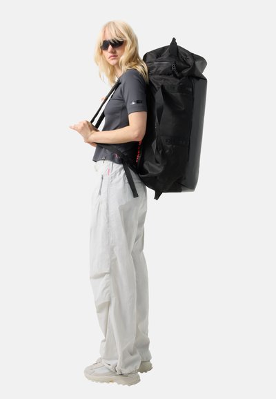 Lyshåret person iført solbriller, sort skjorte, hvide bukser og sneakers, bærende på en stor sort rygsæk, stående i profil.