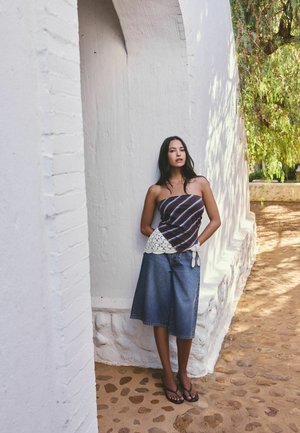 Triibulise rihmadeta topi ja teksapükstega naine seisab valge tekstuurse seina ees munakiviteel, läheduses on rohelust.