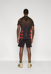 Μαύρη αθλητική ενδυμασία που περιλαμβάνει τοπ με κοντά μανίκια και κόκκινα και χρυσά γραφικά σχέδια, καθώς και ταιριαστά σορτς, συμπληρωμένη με μπεζ παπούτσια.