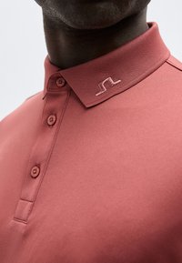 Κοντινό πλάνο ενός ατόμου που φοράει ένα πουκάμισο πόλο σε χρώμα ρόδινη σκόνη με τρία κουμπιά και ένα μικρό κεντημένο λογότυπο στον γιακά.