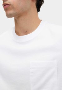 Biała bawełniana koszulka z okrągłym dekoltem, krótkimi rękawami i kieszenią na lewej piersi z płaskim szwem. Gładka tekstura, minimalny design.