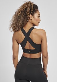 Svart sportbh med korsade band på baksidan, med en enhetlig design och en liten logotypdetalj på den nedre kanten.