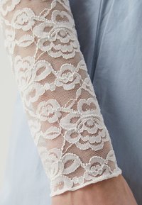 White lace sleeve with floral patterns, transparent texture, and cuff detail against a solid light blue fabric background.