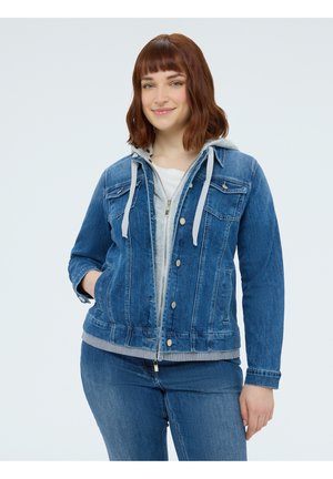 Nuori nainen, jolla on hartioihin ulottuvat ruskeat hiukset, sininen farkkutakki harmaalla hupulla ja siniset farkut, hymyilee ja pitää toista kättään taskussa.