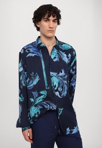 Navyblauwe shirt met een groot turquoise bladpatroon, knoopsluiting aan de voorkant, lange mouwen en een ontspannen pasvorm. Zachte stoftextuur.