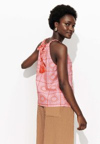 Débardeur rose avec un motif de soleil orange, doté d'un dos noué avec un gland. Associé à un pantalon large beige à rayures. Texture de tissu lisse.