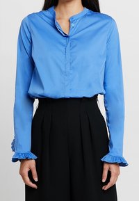 Woman wearing a blue long-sleeve blouse with ruffled cuffs and high-waisted black pleated pants against a plain background.