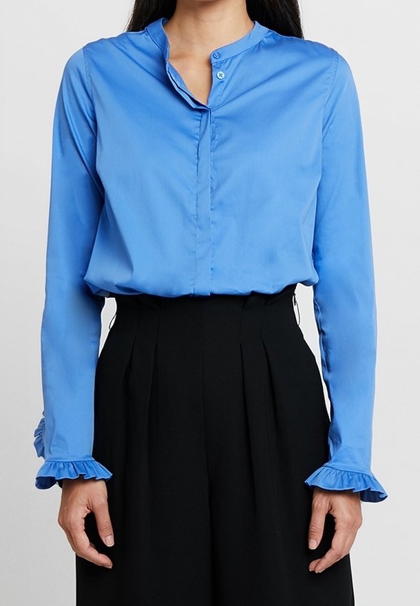 Woman wearing a blue long-sleeve blouse with ruffled cuffs and high-waisted black pleated pants against a plain background.