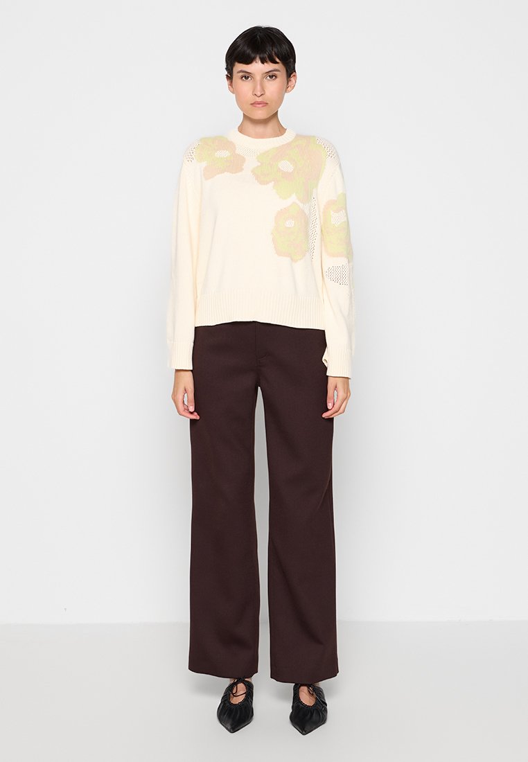 Cremfarbener Pullover mit floralem Muster in hellgrün, kombiniert mit hoch taillierten, dunkelbraunen Hosen und schwarzen, spitz zulaufenden Schuhen.