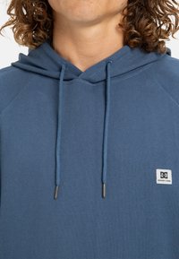El hoodie azul presenta una tela texturizada, cordones ajustables y un pequeño parche con el logo en el lado izquierdo del pecho. Diseñado con un corte relajado.