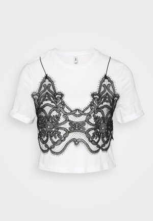 T-shirt blanc à manches courtes ornée d'un overlay en dentelle noire avec des motifs floraux et des bords à volants, créant une esthétique superposée.