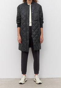 Abrigo largo acolchado negro con cuello alzado, cremallera frontal y bolsillo lateral. Lleva puesto sobre una blusa crema y pantalones oscuros, con zapatillas blancas.