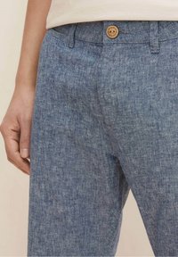 Zbliżenie osoby w niebieskich, teksturowanych lnianych spodniach z guzikiem i widocznymi szlufkami na pasek, oraz w beżowej bluzce częściowo włożonej do środka.