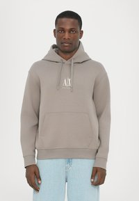 Felpa grigia con cappuccio e tasca frontale; presenta coulisse e un logo bianco sul petto. Indossata con jeans azzurri chiari.