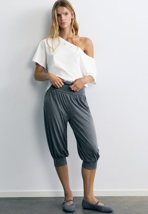 Femme debout près d’un mur, portant un haut blanc épaules dénudées, un pantalon gris ample coupé et des chaussures plates grises, regardant devant elle les mains sur les hanches.