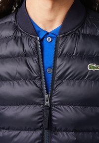 Námořnicky modrá prošívaná péřová bunda s lehkým designem; má zipové zapínání a žebrovaný límec. Pod ní je modré polo triko s knoflíkovými detaily.