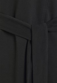 Svart tyg med en mjuk, texturerad yta. Har en bred bälte-detalj knuten vid midjan, vilket skapar en strukturerad silhuett.