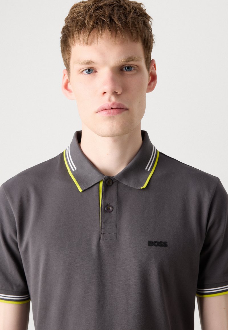 Graues Poloshirt aus weichem Baumwollstoff mit gelben und weißen gestreiften Kragen- und Bündchenakzenten, das ein schwarzes Logo auf der Brust zeigt.