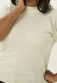 Persoon in een lichtbeige, kortarmige gebreide top met subtiele glans, die de ene hand naar hun nek heft en de andere op hun heup plaatst.