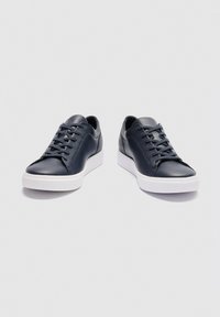 Schwarze flache Ledersneaker mit schwarzen Schnürsenkeln und dicken weißen Gummisohlen auf hellgrauem Hintergrund.