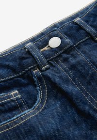 Jeans in denim blu scuro con un bottone in metallo argentato e cuciture gialle a contrasto, con una visione ravvicinata della vita e dei dettagli della tasca.