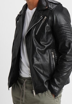 Homme portant une veste de motard en cuir noir avec des poches zippées, un t-shirt blanc et un pantalon vert à cordon, les mains dans les poches.