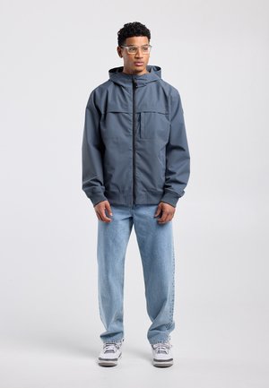 Man draagt een transparante bril, een blauwe capuchonvest met rits, lichtblauwe spijkerbroek en witte sneakers, staat ontspannen tegen een effen achtergrond.