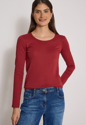 Donna con capelli lunghi e scuri che indossa una maglietta rossa a maniche lunghe e jeans blu, sorridente con una mano sul fianco.